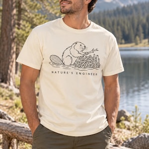 Może przedstawiać: Kremowy t-shirt z krótkim rękawem z czarną grafiką liniową przedstawiającą bobra budującego tamę z patyków. Poniżej ilustracji znajduje się napis "NATURE'S ENGINEER" wykonany wyraźną, bezszeryfową czcionką na środku klatki piersiowej.