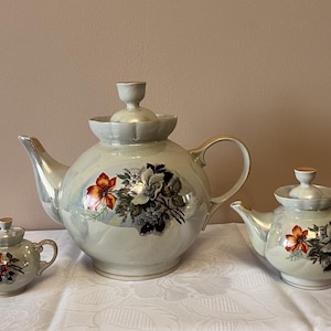 Könnte beinhalten: Ein Set aus drei Keramik-Teekannen in verschiedenen Größen. Die Teekannen sind perlmuttweiß mit goldenen Akzenten und floralen Designs in Rot, Orange und Schwarz. Das Set enthält eine große Teekanne und zwei kleinere.