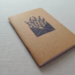 May include: A small brown notebook with a white spine. The cover has a black ink stamp of a bouquet of flowers in an envelope.