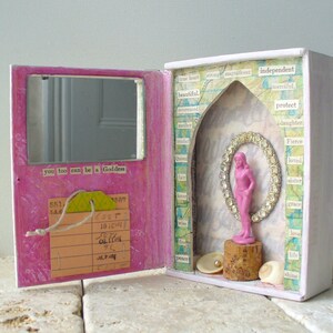 May include: A handmade pink and white box with a mirror and a small pink figurine inside. The box has a hinged door with a mirror and the text "you too can be a Goddess". Inside, a pink figurine stands on a cork.
