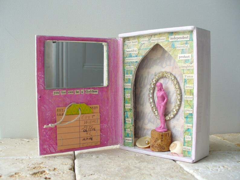 May include: A handmade pink and white box with a mirror and a small pink figurine inside. The box has a hinged door with a mirror and the text "you too can be a Goddess". Inside, a pink figurine stands on a cork.