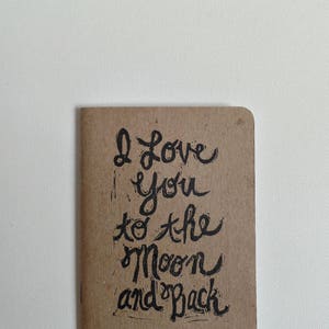 May include: A brown paper notebook with the handwritten message "I love you to the moon and back" in black ink.