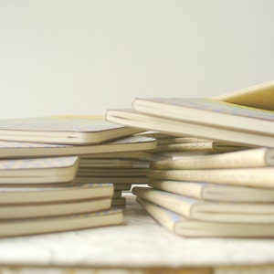 May include: A stack of notebooks with a variety of patterned covers. The top notebook has a light blue and white patterned cover. The notebooks are stacked on a white surface.