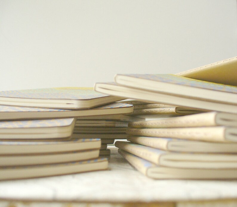 May include: A stack of notebooks with a variety of patterned covers. The top notebook has a light blue and white patterned cover. The notebooks are stacked on a white surface.