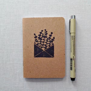 May include: A small brown notebook with a black ink stamp of a plant in an envelope. The notebook is next to a beige pen on a white surface.