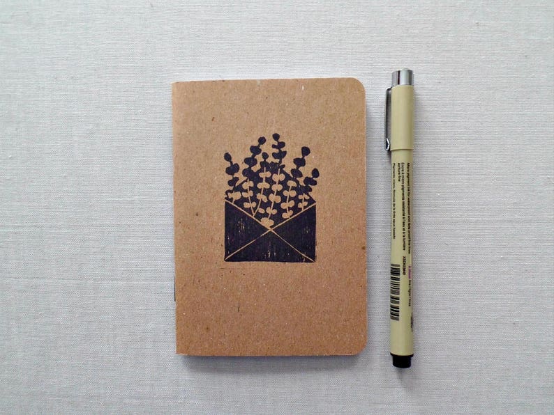 May include: A small brown notebook with a black ink stamp of a plant in an envelope. The notebook is next to a beige pen on a white surface.