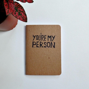 Op de afbeelding: Een klein bruin notitieboekje met de woorden "You're my person" geschreven in zwarte inkt.