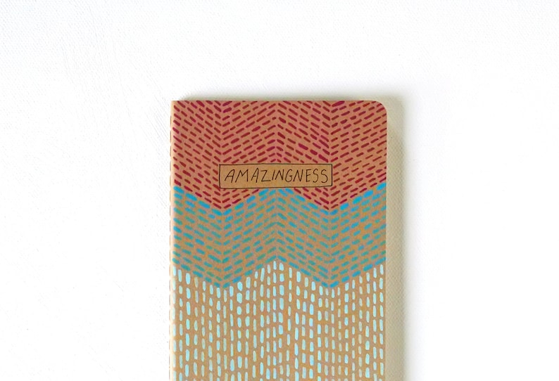 May include: A small notebook with a brown cover and a colorful geometric pattern. The cover has three sections: a red and orange chevron pattern, a blue and turquoise chevron pattern, and a light blue and white polka dot pattern. The notebook has a brown label with the word "AMAZINGNESS" written in black.