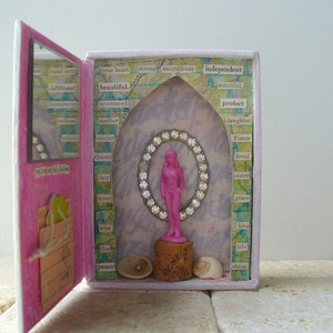 May include: A mixed media art piece featuring a pink box with an open door. Inside, a pink figurine is framed by a jeweled oval. The box is decorated with words like "beautiful" and "independent". Seashells and a cork base are at the bottom.