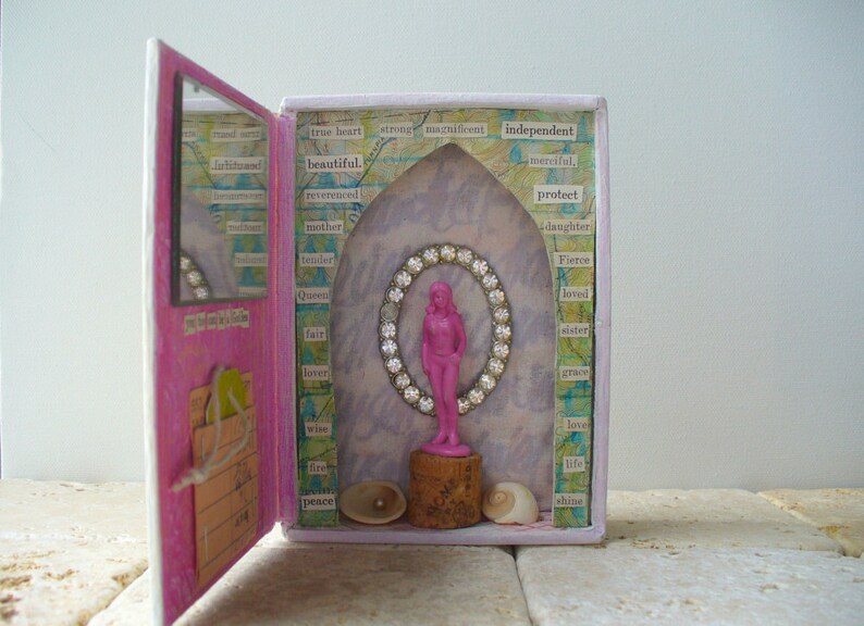 May include: A mixed media art piece featuring a pink box with an open door. Inside, a pink figurine is framed by a jeweled oval. The box is decorated with words like "beautiful" and "independent". Seashells and a cork base are at the bottom.