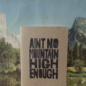 K&ouml;nnte beinhalten: Ein braunes Notizbuch mit dem Text "Ain't no mountain high enough" in Schwarz gedruckt.