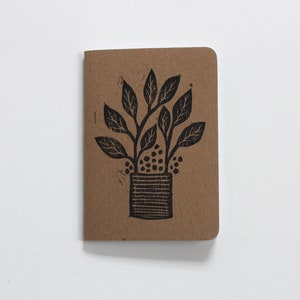 May include: A brown notebook featuring a black linocut illustration of a potted plant with leaves and berries.