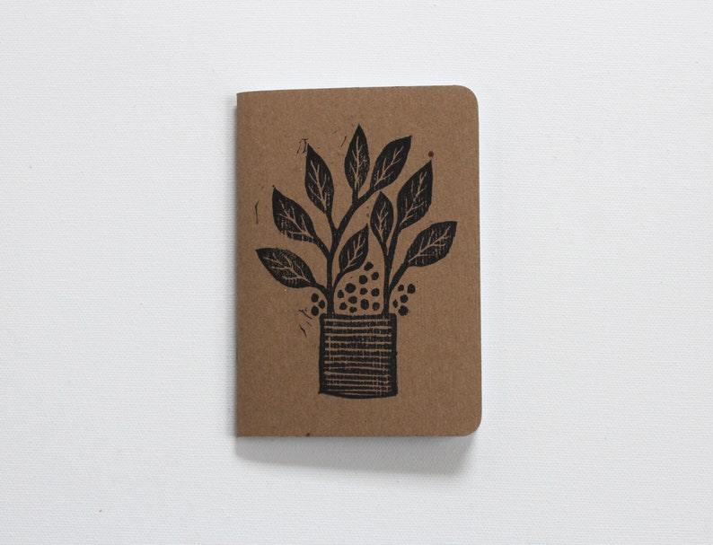 May include: A brown notebook featuring a black linocut illustration of a potted plant with leaves and berries.
