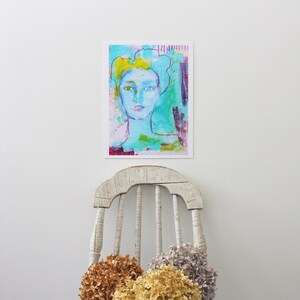 May include: An abstract art print featuring a portrait of a person with blue skin and yellow hair. The artwork uses a variety of colors, including turquoise, pink, and purple. The print is displayed on a white wall above a distressed white wooden chair.