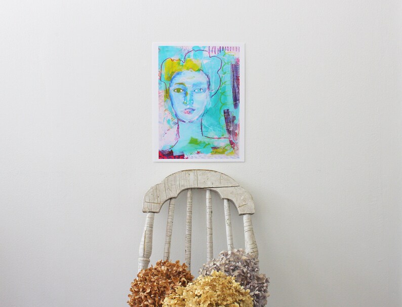 May include: An abstract art print featuring a portrait of a person with blue skin and yellow hair. The artwork uses a variety of colors, including turquoise, pink, and purple. The print is displayed on a white wall above a distressed white wooden chair.