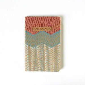 May include: A small, brown notebook with a red, blue, and white geometric pattern on the cover. The cover has the word "AMAZINGNESS" printed on it in a brown rectangle.