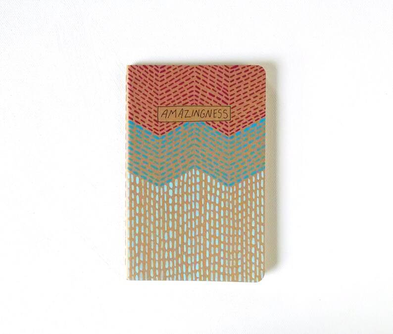 May include: A small, brown notebook with a red, blue, and white geometric pattern on the cover. The cover has the word "AMAZINGNESS" printed on it in a brown rectangle.
