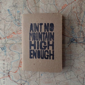 K&ouml;nnte beinhalten: Ein braunes Notizbuch mit dem Text "Ain't No Mountain High Enough" in Schwarz gedruckt.