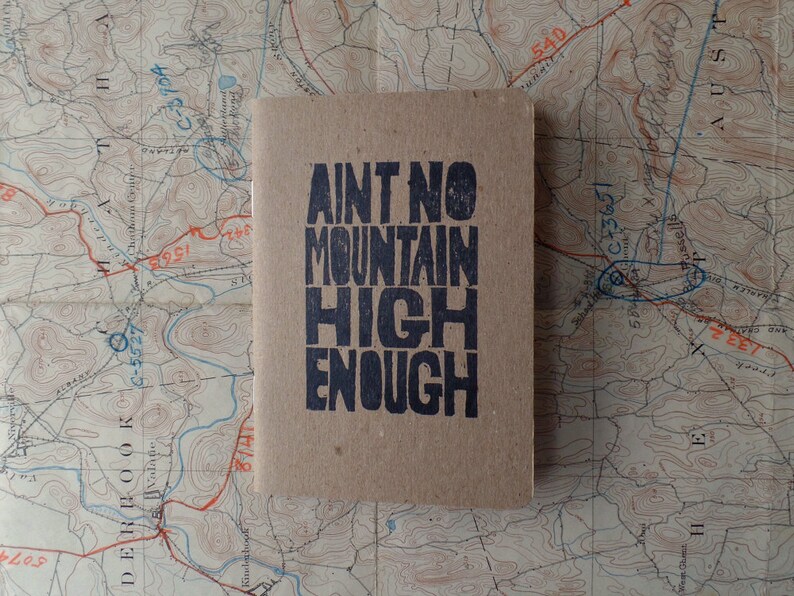 K&ouml;nnte beinhalten: Ein braunes Notizbuch mit dem Text "Ain't No Mountain High Enough" in Schwarz gedruckt.