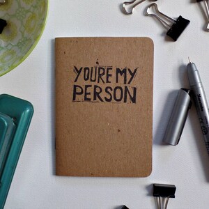 Op de afbeelding: Een klein bruin notitieboekje met de woorden "YOU'RE MY PERSON" in zwarte inkt op de voorkant.