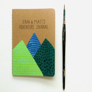May include: A brown journal with the text "Erin & Matt's Adventure Journal" on the front cover. The cover is decorated with three green, blue, and light green mountains with different patterns.