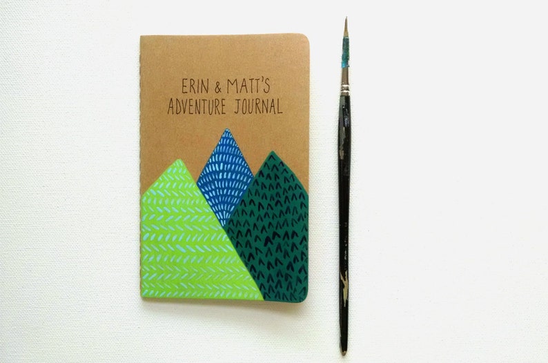 May include: A brown journal with the text "Erin & Matt's Adventure Journal" on the front cover. The cover is decorated with three green, blue, and light green mountains with different patterns.