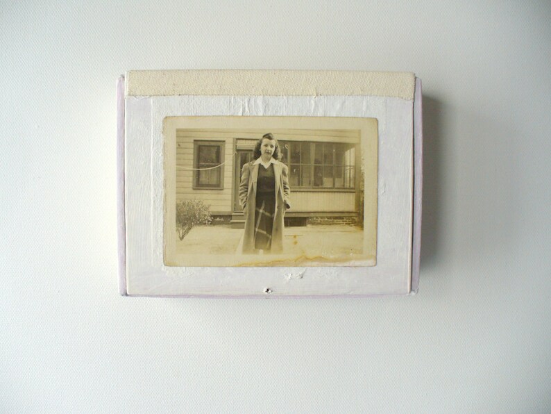 May include: A vintage black and white photograph of a woman standing in front of a house, framed in a white and lavender box. The woman is wearing a long coat and dress. The photograph is slightly faded.