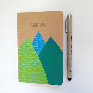 May include: A brown notebook with a green, blue, and teal mountain design and the word "Adventures" written on the front.