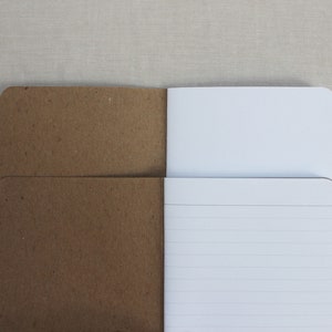 May include: A brown paper notebook with a white lined interior. The notebook is open to reveal the lined pages.