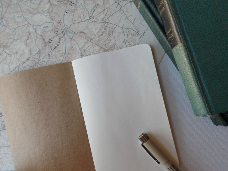 May include: An open notebook with a brown cover and blank white pages. A beige pen is lying on the open pages. The notebook is on a white surface with a map and a stack of green books in the background.