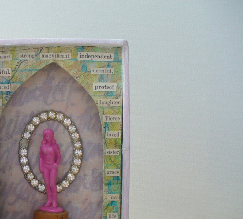 May include: A mixed media artwork featuring a pink figurine within a jeweled oval frame. The background includes words like "independent" and "fierce" on a textured surface. The artwork is framed in a light purple border.