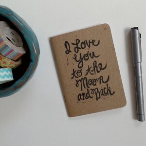 May include: A brown card with the handwritten message "I love you to the moon and back" on a white surface with a silver pen.