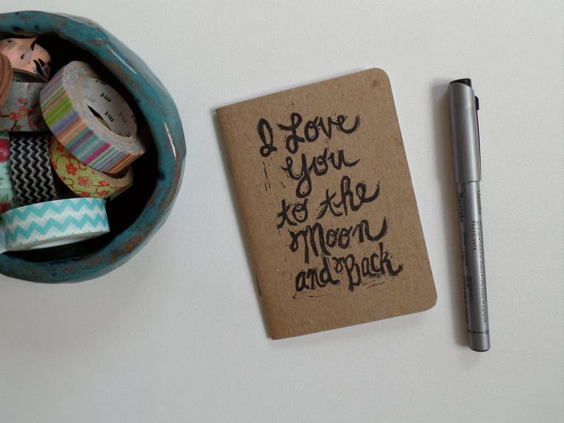 May include: A brown card with the handwritten message "I love you to the moon and back" on a white surface with a silver pen.