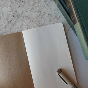May include: An open notebook with a brown cover and blank white pages. A beige pen is lying on the open pages. The notebook is on a white surface with a map and a stack of green books in the background.