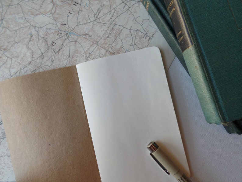May include: An open notebook with a brown cover and blank white pages. A beige pen is lying on the open pages. The notebook is on a white surface with a map and a stack of green books in the background.