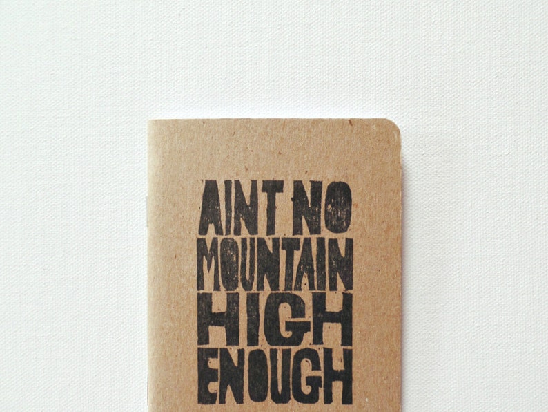 K&ouml;nnte beinhalten: Ein braunes Notizbuch mit dem Text "Ain't no mountain high enough" in Schwarz gedruckt.