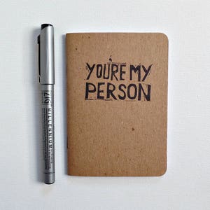 Op de afbeelding: Een klein bruin notitieboekje met de woorden "You're my person" in zwarte inkt op de voorkant.