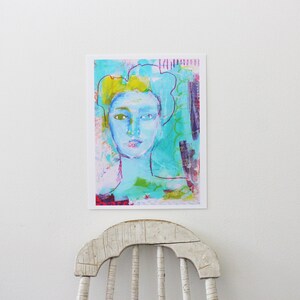 May include: Abstract art print featuring a portrait of a person with a light blue face and yellow hair. The artwork uses a mix of colors, including turquoise, pink, and purple. The art print is displayed on a white wall.