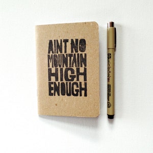 K&ouml;nnte beinhalten: Ein kleines braunes Notizbuch mit dem Text "Ain't no mountain high enough" auf dem vorderen Umschlag. Neben dem Notizbuch liegt ein brauner Stift.