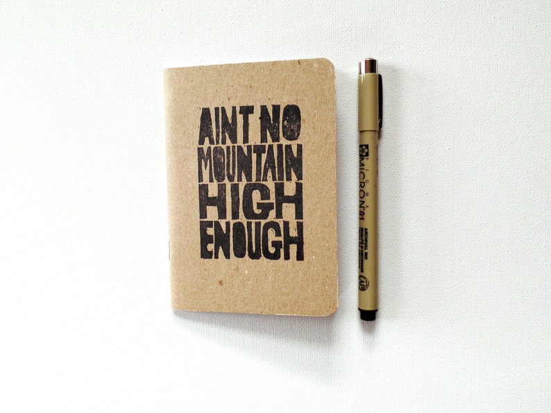 K&ouml;nnte beinhalten: Ein kleines braunes Notizbuch mit dem Text "Ain't no mountain high enough" auf dem vorderen Umschlag. Neben dem Notizbuch liegt ein brauner Stift.