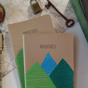 May include: Two brown notebooks with the word "Adventures" written on the front. The top notebook has a green and blue mountain design on the front.