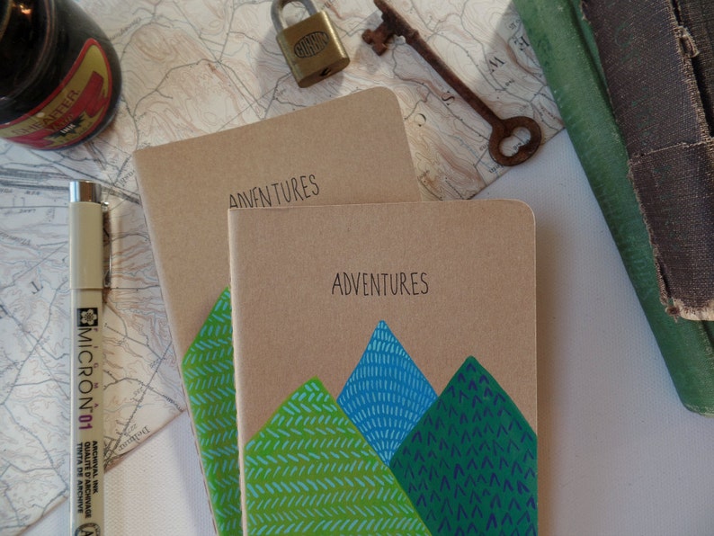 May include: Two brown notebooks with the word "Adventures" written on the front. The top notebook has a green and blue mountain design on the front.