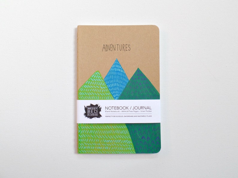 May include: A brown notebook with a green, blue, and white mountain illustration on the cover. The notebook says "Adventures" and "Notebook/Journal" on the front. The notebook is blank, has 64 acid-free pages, and an inner pocket.