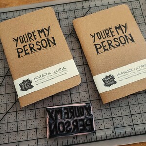 Op de afbeelding: Twee bruine notitieboekjes met de tekst "You're My Person" gedrukt op de voorkant. De notitieboekjes liggen op een zwart-witte snijmat. Een rubberen stempel met dezelfde tekst bevindt zich op de voorgrond.