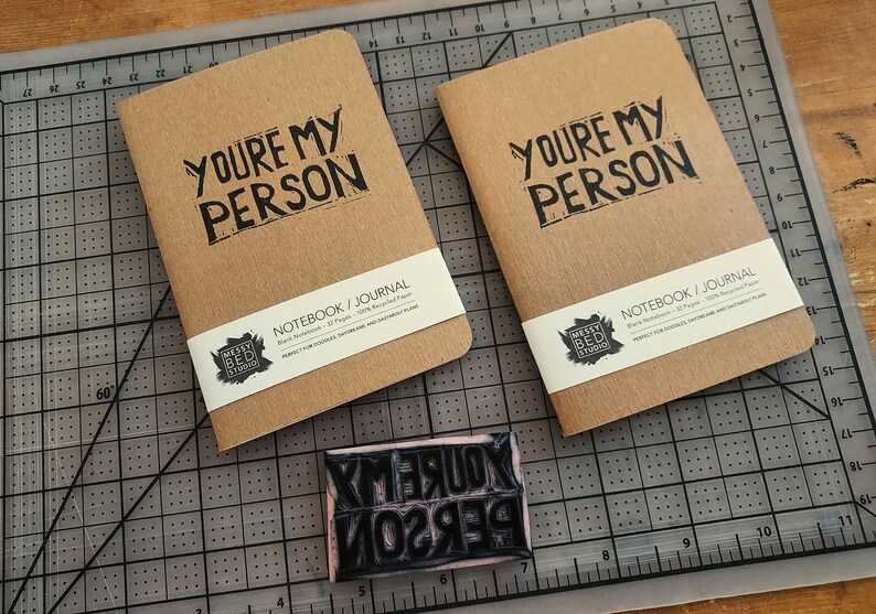 Op de afbeelding: Twee bruine notitieboekjes met de tekst "You're My Person" gedrukt op de voorkant. De notitieboekjes liggen op een zwart-witte snijmat. Een rubberen stempel met dezelfde tekst bevindt zich op de voorgrond.