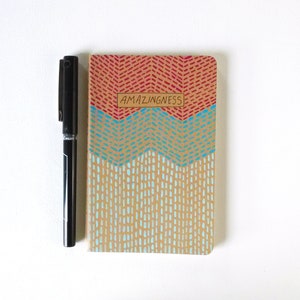 May include: A small, brown notebook with a red, blue, and beige geometric pattern. The notebook has the word "AMAZINGNESS" written on the front in black.