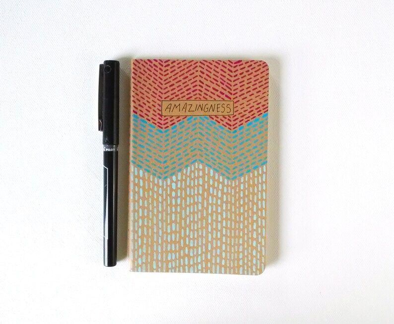 May include: A small, brown notebook with a red, blue, and beige geometric pattern. The notebook has the word "AMAZINGNESS" written on the front in black.