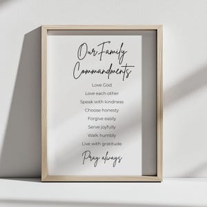 Può includere: Una stampa incorniciata con la scritta "Our Family Commandments" in elegante corsivo. La stampa elenca comandamenti come "Ama Dio", "Amatevi gli uni gli altri" e "Pregate sempre". La cornice è in legno chiaro e lo sfondo è bianco.