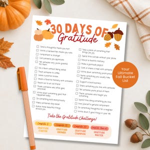 May include: A white paper sheet titled 30 Days of Gratitude with a list of daily tasks, decorated with orange pumpkins, brown acorns, and autumn leaves. The text includes a challenge section and an orange circle stating Your Ultimate Fall Bucket List.