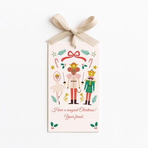 Könnte beinhalten: Ein rosa Geschenkanhänger mit beigem Band zeigt eine festliche Illustration. Das Design umfasst eine Ballerina, einen Mäusekönig und einen Nussknackersoldaten mit Weihnachtselementen wie Zuckerstangen und Stechpalmen. Der Text lautet: "Frohe magische Weihnachten! Dein Freund."